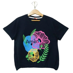 A black short-sleeve t-shirt hanging on a wooden hanger, featuring a colorful graphic print with a pink flower, a yellow flower, green leaves, and a blue circle containing the word 'Amen'.