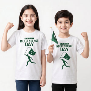Two children modeling white t-shirts with a green 'HAPPY INDEPENDENCE DAY' graphic and a running figure holding a Pakistan flag. The girl on the left raises her fist, and the boy on the right holds a small Pakistan flag against a plain white background.