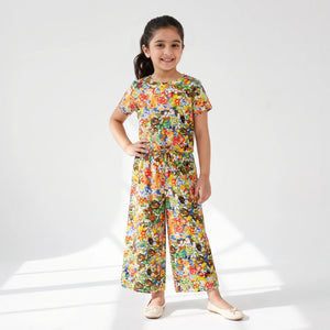 A young girl stands in a studio wearing a colorful short-sleeve jumpsuit with a dense floral and abstract print. The jumpsuit features a round neckline, short sleeves, a matching waist tie, and wide legs.