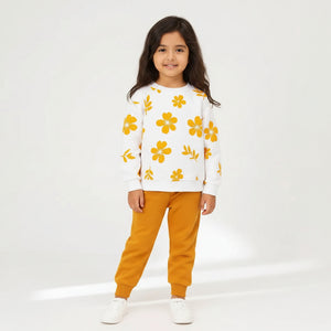 A young girl modeling a white crewneck sweatshirt with a yellow floral pattern and matching mustard yellow jogger pants, standing against a plain off-white background.
