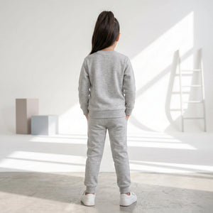 A child standing with their back to the camera, wearing a light gray heathered sweatshirt and matching jogger pants with white sneakers, in a bright, modern room with geometric blocks and a ladder.