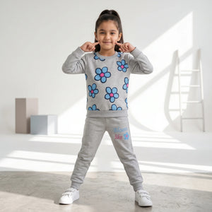 A young girl stands in a bright, modern room wearing a light gray two-piece sweatshirt and jogger set. The sweatshirt features a pattern of blue flowers with pink centers, and the joggers have 'Moon Lake' printed on the thigh.