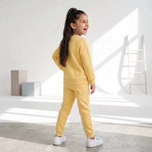 A young girl with long dark hair in a ponytail is shown from the back wearing a matching yellow sweatshirt and jogger pants set, paired with white sneakers, in a bright, modern minimalist room with sunlight and shadows.