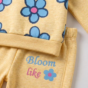 A close-up of a children's two-piece outfit in heathered pale yellow, featuring a long-sleeve top with blue flowers and the pants printed with the words 'Bloom like' in blue and pink alongside a small flower graphic.