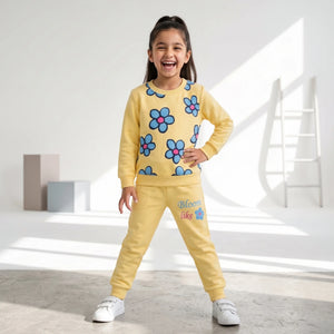 A young girl smiling and posing in a yellow two-piece pajama set with blue and pink flower prints, featuring the text 'Bloom like a flower' on the pants, set against a bright, modern white studio background with geometric props.