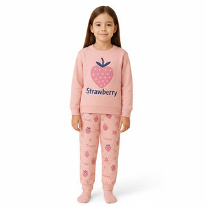 A young girl modeling a pink two-piece pajama set consisting of a sweatshirt with a strawberry graphic and the word 'Strawberry' printed on the chest, and matching patterned pants, against a solid light beige background.