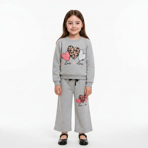 A young girl stands indoors wearing a light gray two-piece sweatshirt and jogger pants set with heart and leopard-print graphic details. The background shows a neutral wall and wooden staircase.