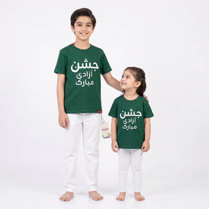 A boy and a girl standing barefoot against a plain white studio background, wearing matching dark green T-shirts with white Arabic text and white pants.