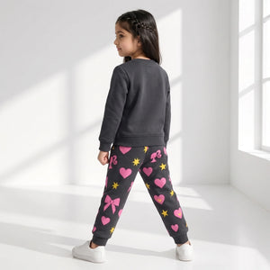 A young girl is shown from the back wearing a dark gray sweatshirt and matching dark gray joggers with pink hearts, bows, and yellow stars, standing in a bright white room with window light.