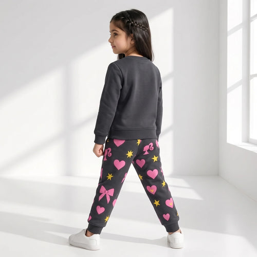 A young girl is shown from the back wearing a dark gray sweatshirt and matching dark gray joggers with pink hearts, bows, and yellow stars, standing in a bright white room with window light.