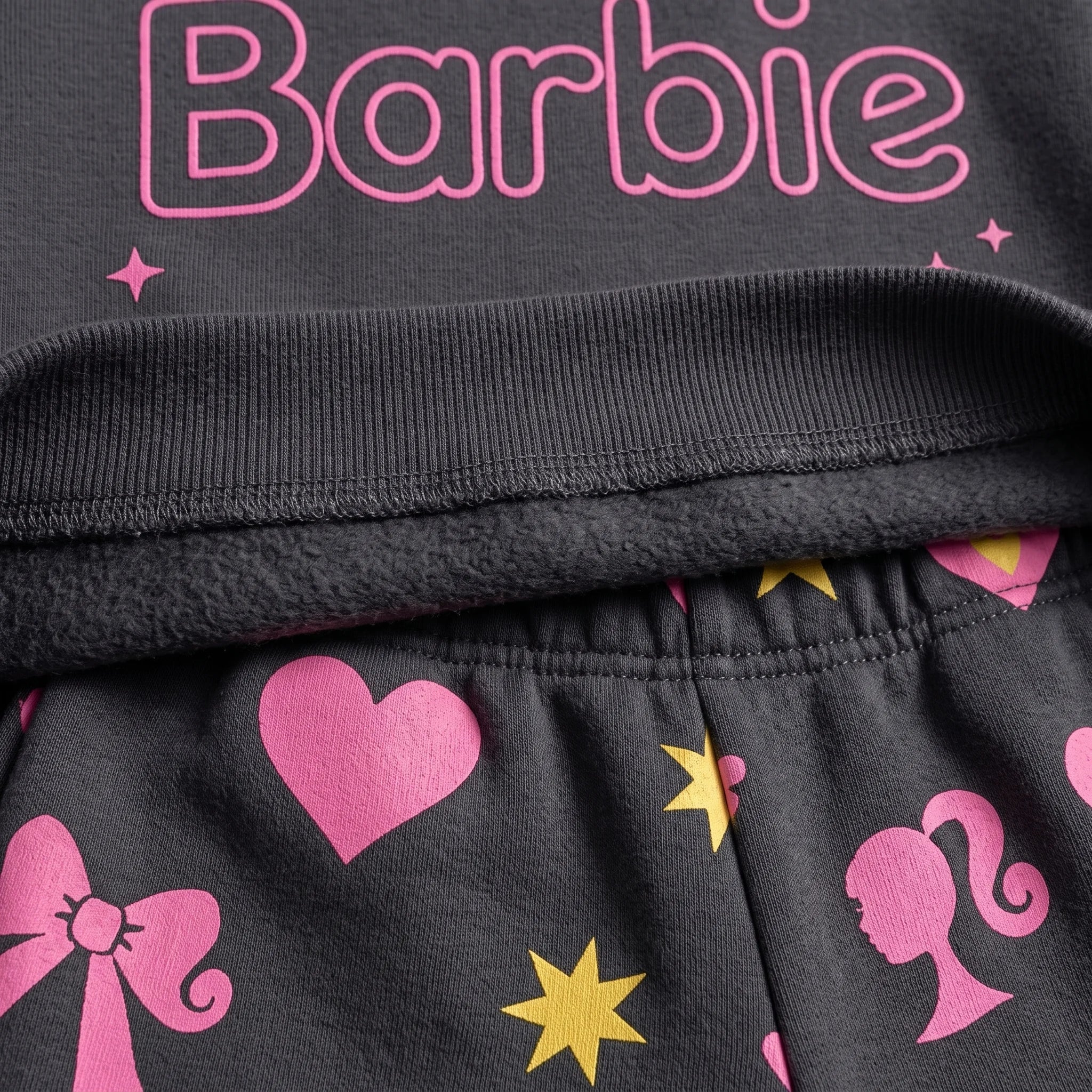 Close-up of a black children's garment featuring pink Barbie text across the chest, a pink heart, yellow star, and pink silhouette motifs on the fabric below the ribbed waistband.
