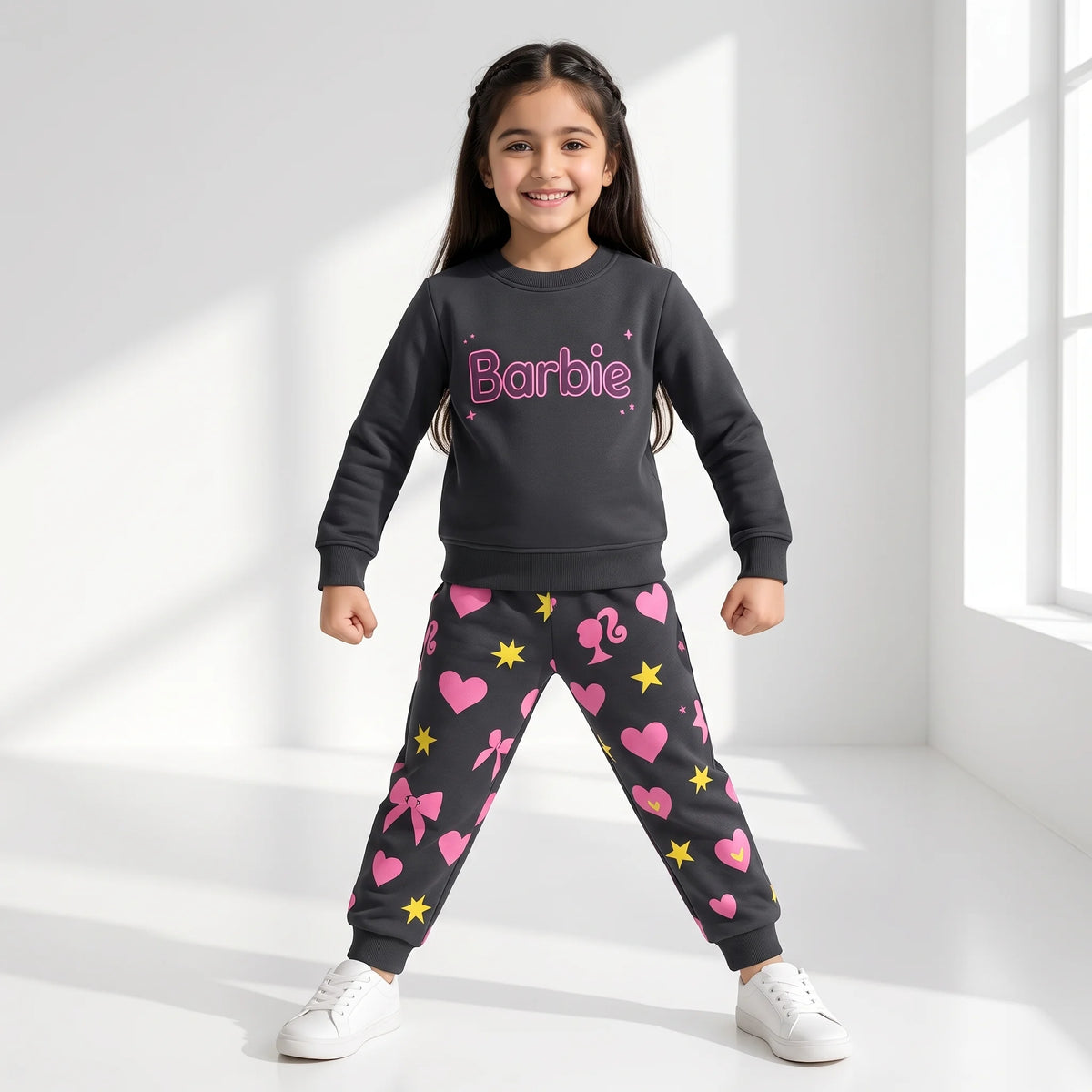 A young girl stands in a bright white room wearing a dark gray sweatshirt and matching jogger pants with pink hearts, bows, stars, and the Barbie logo on the chest.