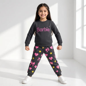 A young girl stands in a bright white room wearing a dark gray sweatshirt and matching jogger pants with pink hearts, bows, stars, and the Barbie logo on the chest.