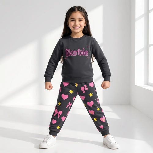 A young girl stands in a bright white room wearing a dark gray sweatshirt and matching jogger pants with pink hearts, bows, stars, and the Barbie logo on the chest.