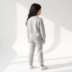 A child model shown from the back wearing a light gray heather sweatshirt and matching jogger pants with white sneakers, standing against a white studio wall with soft geometric shadows.