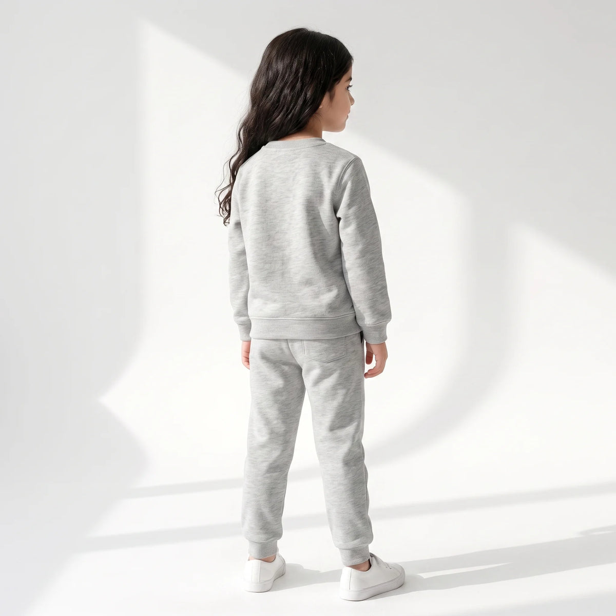 A child model shown from the back wearing a light gray heather sweatshirt and matching jogger pants with white sneakers, standing against a white studio wall with soft geometric shadows.