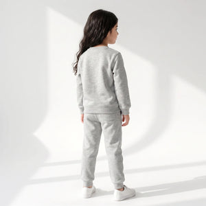 A child model shown from the back wearing a light gray heather sweatshirt and matching jogger pants with white sneakers, standing against a white studio wall with soft geometric shadows.
