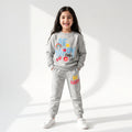 A young girl stands smiling in a studio setting wearing a light gray sweatshirt and matching jogger pants with colorful 'BE HAPPY EVERY DAY' graphics on the front and 'BE HAPPY' on the thigh.