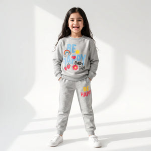 A young girl stands smiling in a studio setting wearing a light gray sweatshirt and matching jogger pants with colorful 'BE HAPPY EVERY DAY' graphics on the front and 'BE HAPPY' on the thigh.