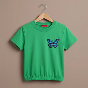 A bright green short-sleeve kids' t-shirt with a blue and black butterfly graphic on the left chest, hanging on a wooden hanger against a solid taupe background.