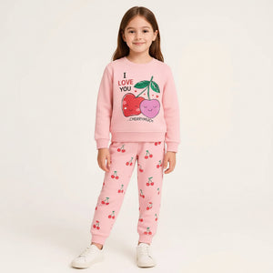A young girl modeling a pink two-piece sweatshirt and jogger set with a cherry graphic on the sweatshirt and a cherry print on the pants, set against a plain light background.