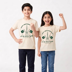 A boy and a girl modeling matching cream-colored t-shirts with a green and black 'Happy Independence Day' graphic featuring the Pakistan flag and the Arqel Tower. The boy wears dark navy pants, and the girl wears blue jeans, standing against a plain white studio background.