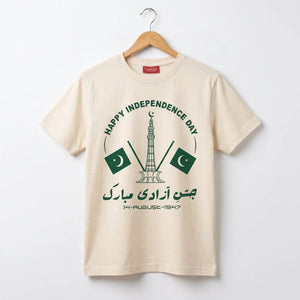A beige short-sleeve t-shirt hanging on a wooden hanger against a light gray wall. The shirt features a dark green graphic on the front with the text 'HAPPY INDEPENDENCE DAY', a mosque tower, two Pakistani flags, and Urdu text 'جمن آبادی مہرل 14-AUGUST-1947'.