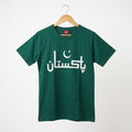 A dark green short-sleeve t-shirt hanging on a wooden hanger against a light gray background. The shirt features the word 'Pakistan' printed in white Arabic calligraphy across the chest, with a white crescent and star graphic integrated above the letters.