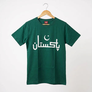 A dark green short-sleeve t-shirt hanging on a wooden hanger against a light gray background. The shirt features the word 'Pakistan' printed in white Arabic calligraphy across the chest, with a white crescent and star graphic integrated above the letters.