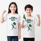 Two children modeling white t-shirts with a green 'HAPPY INDEPENDENCE DAY' graphic and a running figure holding a Pakistan flag. The girl on the left raises her fist, and the boy on the right holds a small Pakistan flag against a plain white background.