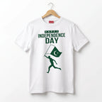 A white short-sleeve t-shirt hanging on a wooden hanger against a plain white background. The shirt features a green graphic print that reads 'HAPPY INDEPENDENCE DAY' above a silhouette of a runner carrying a Pakistan flag.
