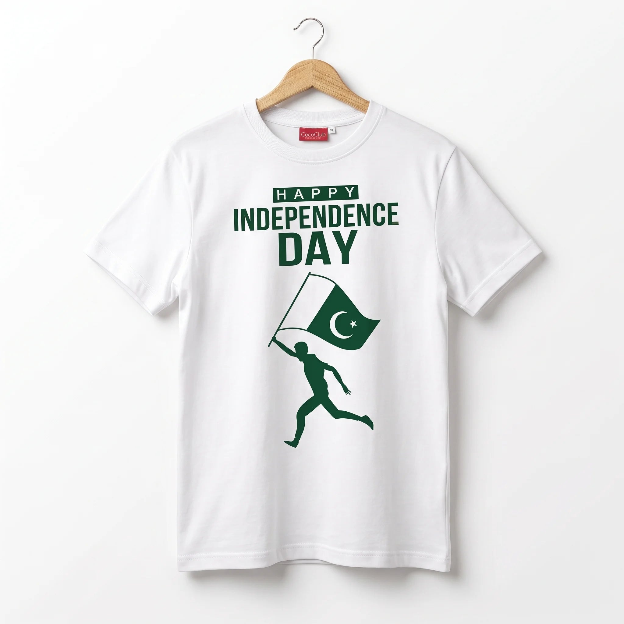 A white short-sleeve t-shirt hanging on a wooden hanger against a plain white background. The shirt features a green graphic print that reads 'HAPPY INDEPENDENCE DAY' above a silhouette of a runner carrying a Pakistan flag.