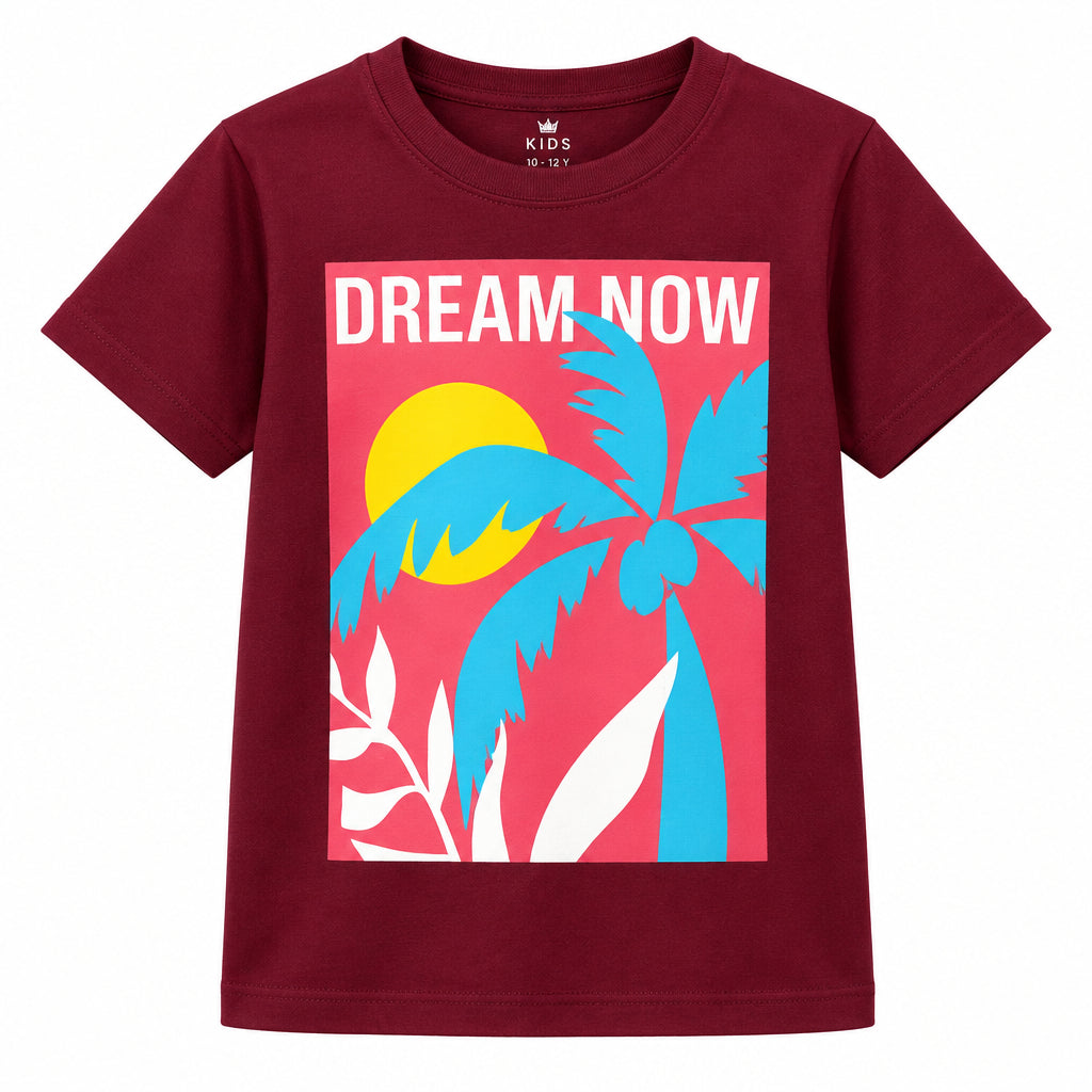 A young girl stands in a bright living room wearing a maroon graphic t-shirt with a colorful tropical design and the words 'DREAM NOW', paired with blue jeans and white sneakers.