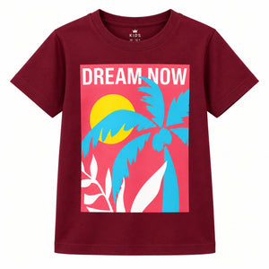 A young girl stands in a bright living room wearing a maroon graphic t-shirt with a colorful tropical design and the words 'DREAM NOW', paired with blue jeans and white sneakers.