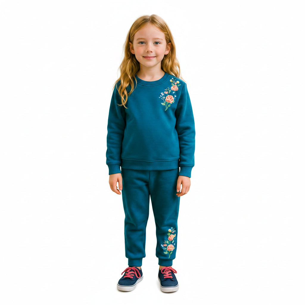 A young girl with long blonde hair stands in a living room wearing a matching teal blue sweatshirt and jogger pants set with colorful floral embroidery on the chest and left thigh.