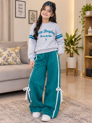 A young girl stands in a living room wearing a light gray sweatshirt with blue 'Los Angeles' lettering and teal wide-leg pants with white side stripes and tie details.