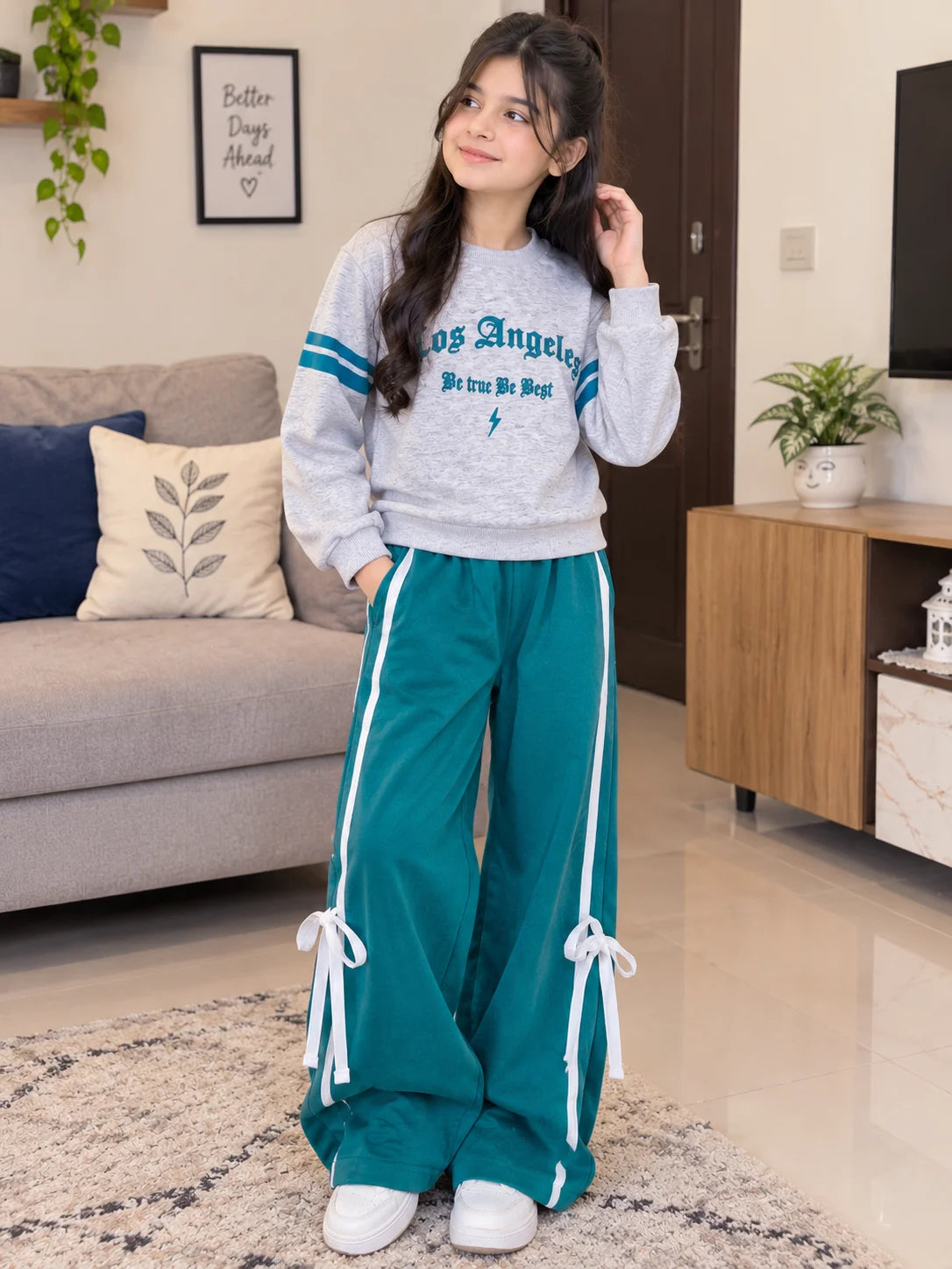 A young woman stands in a living room wearing a light gray sweatshirt with teal lettering and stripes, paired with teal wide-leg pants featuring white side stripes and tie details at the hems.