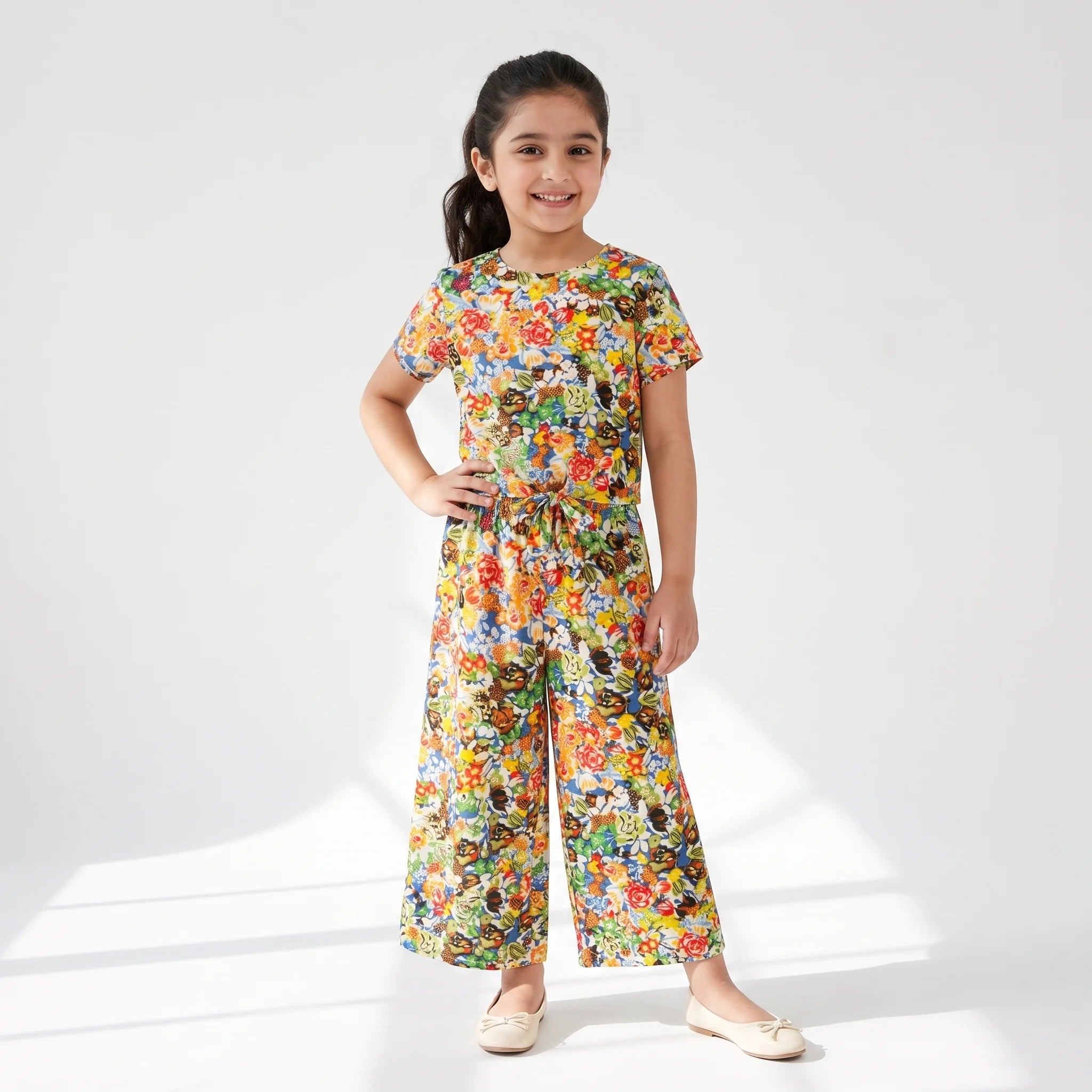 A young girl stands in a studio wearing a colorful short-sleeve jumpsuit with a dense floral and abstract print. The jumpsuit features a round neckline, short sleeves, a matching waist tie, and wide legs.