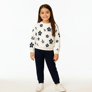 Young girl wearing a white sweater with black floral patterns and dark blue pants in a living room setting.