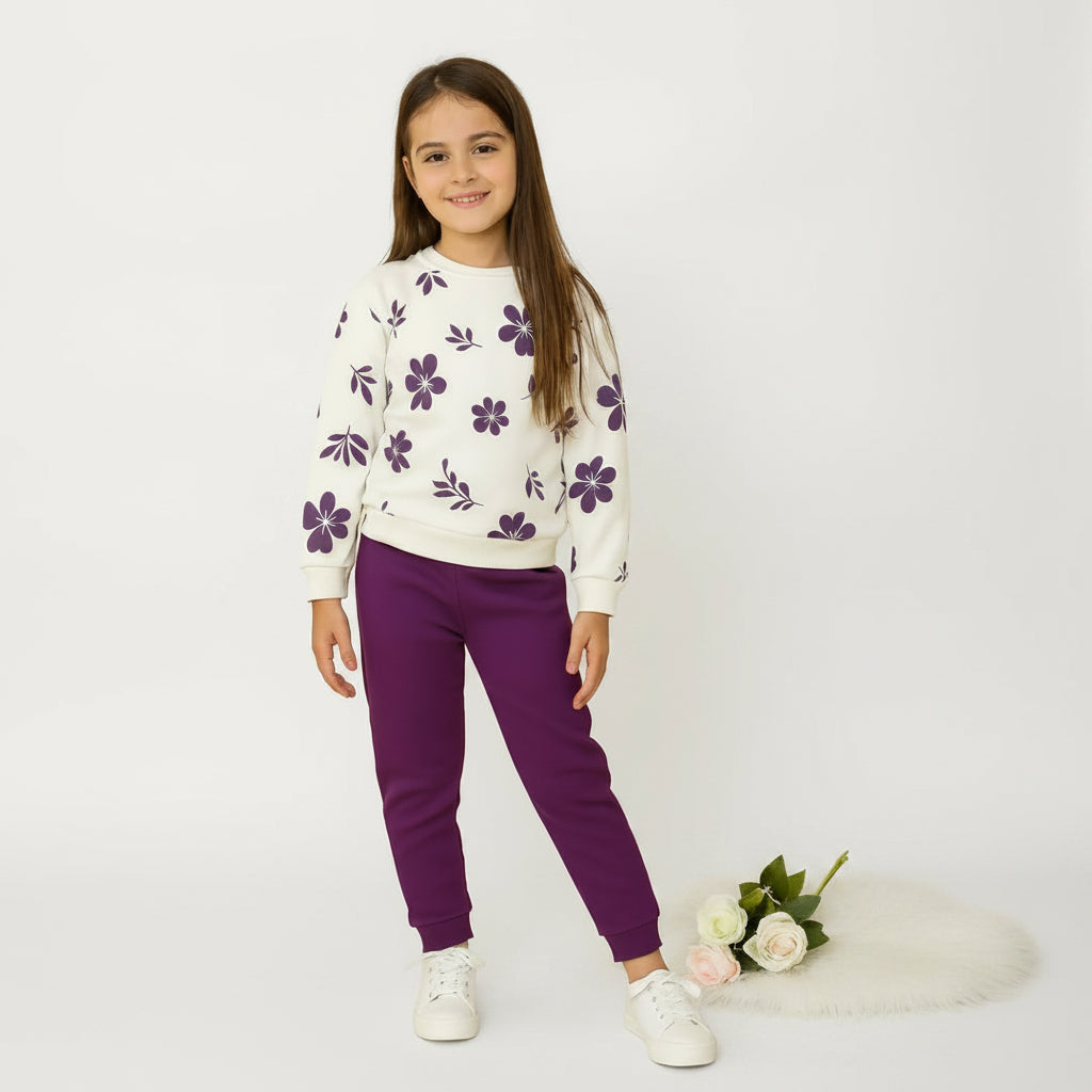 A young girl stands against a warm beige background wearing a white crewneck sweatshirt with purple floral patterns, matching purple jogger pants, and white sneakers. A small bouquet of pink and white roses rests on a white faux-fur rug near her right side.