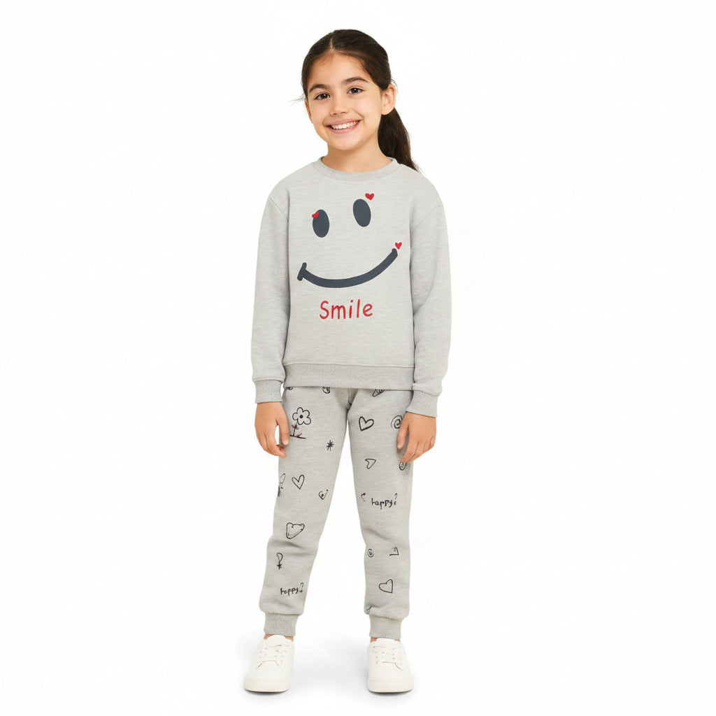 A young girl stands against a light cream background wearing a light gray two-piece sweatshirt and jogger pants set. The sweatshirt features a large black smiley face with small red heart accents and the word 'Smile' in red.