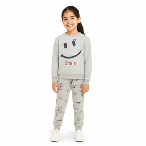 A young girl stands against a light cream background wearing a light gray two-piece sweatshirt and jogger pants set. The sweatshirt features a large black smiley face with small red heart accents and the word 'Smile' in red.