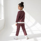 A young girl with her hair in a bun is wearing a matching two-piece tracksuit in a dusty plum-brown color with white ribbed trim, standing in a bright white room with window shadows.