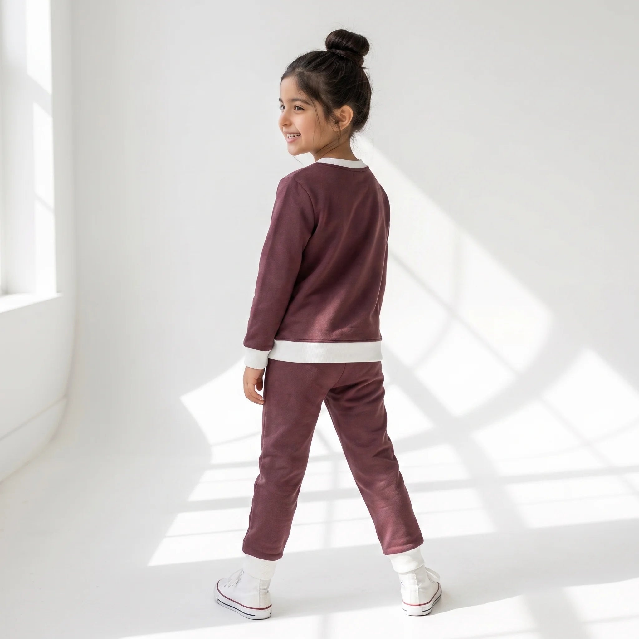 A young girl with her hair in a bun is wearing a matching two-piece tracksuit in a dusty plum-brown color with white ribbed trim, standing in a bright white room with window shadows.