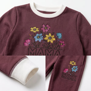 A child's crewneck sweatshirt in maroon with a white ribbed collar and cuffs, featuring a colorful embroidered floral design and the word 'MAMA' on the front. A close-up inset shows the white ribbed cuff, and another inset shows the matching sweatpants leg with the embroidery.