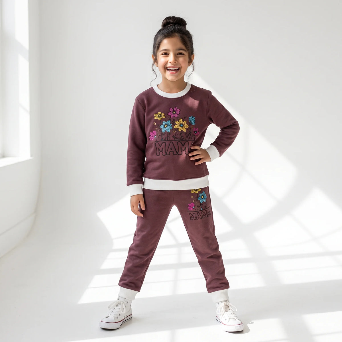 A young girl smiling in a matching maroon sweatshirt and jogger pants set with white trim and colorful floral graphics that read 'MANY MANY MAMAS'.