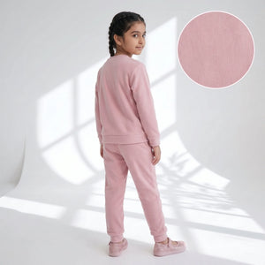 A young girl with dark braided hair is shown from the back wearing a light pink two-piece lounge set consisting of a long-sleeve sweatshirt and matching jogger pants. She is standing in a bright white studio with window shadow projections, and a circular fabric texture inset is visible in the top right corner.