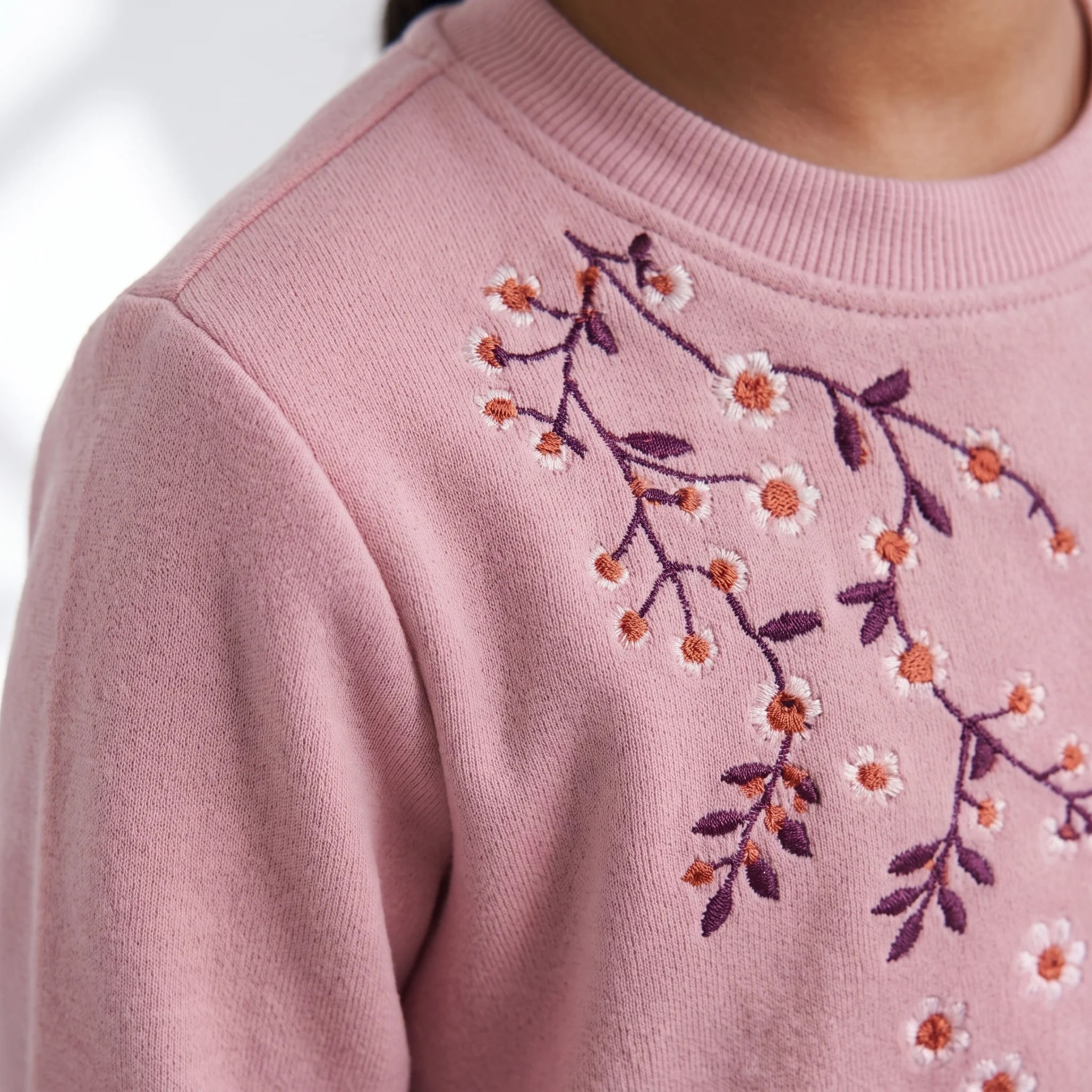 Close-up of a child wearing a dusty pink crewneck sweatshirt with embroidered cherry blossom branches in white, orange, and purple on the chest.