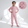 A young girl modeling a light pink two-piece sweatshirt and jogger pants set with floral embroidery, standing against a white studio backdrop with window shadow projections. An inset circle in the upper right shows a close-up of the embroidery detail.