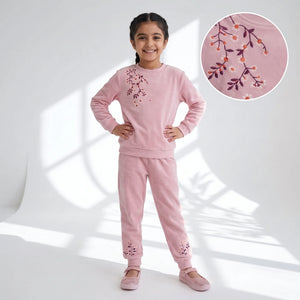 A young girl modeling a light pink two-piece sweatshirt and jogger pants set with floral embroidery, standing against a white studio backdrop with window shadow projections. An inset circle in the upper right shows a close-up of the embroidery detail.